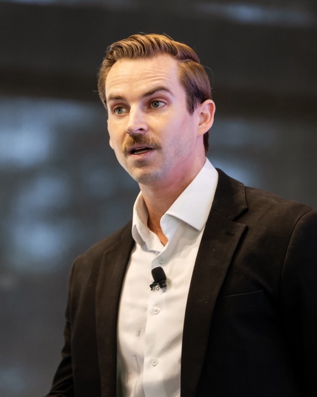 Lucas Miller speaking, mid-sentence, in a dark blazer and white shirt